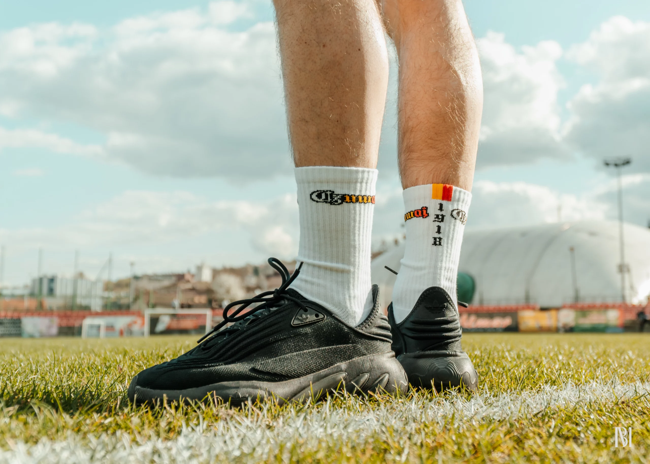 Białe skarpetki z mini flagą o kolorach biało-żółto-czerwonych i rokiem 1918 na tle stadionu, krzesełek i budynku klubowego.