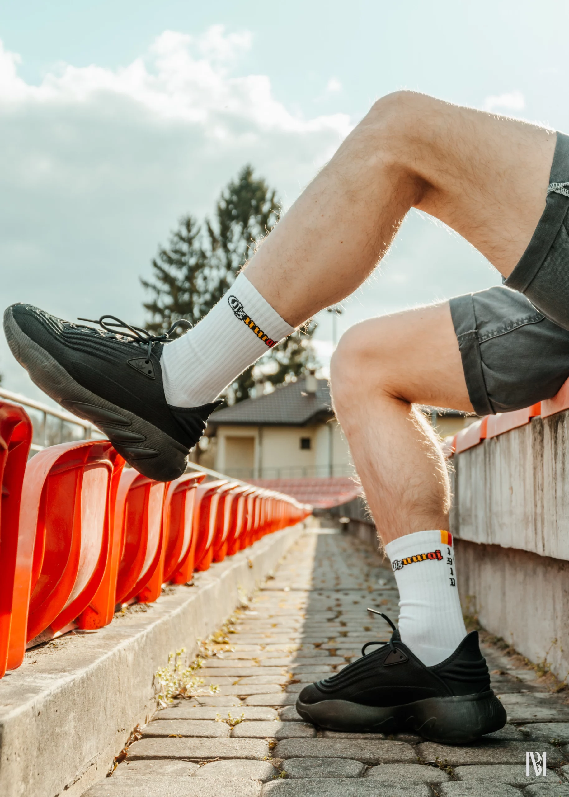 Białe skarpetki z mini flagą o kolorach biało-żółto-czerwonych i rokiem 1918 na tle stadionu, krzesełek i budynku klubowego.
