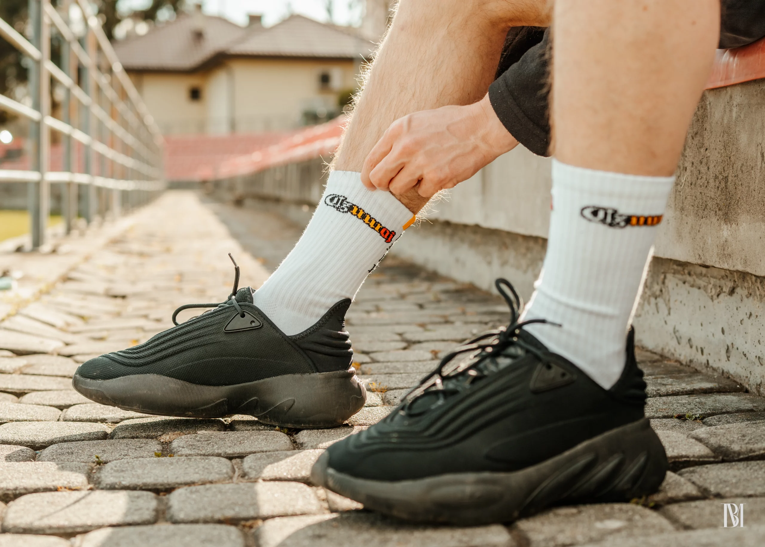 Białe skarpetki z mini flagą o kolorach biało-żółto-czerwonych i rokiem 1918 na tle stadionu, krzesełek i budynku klubowego.