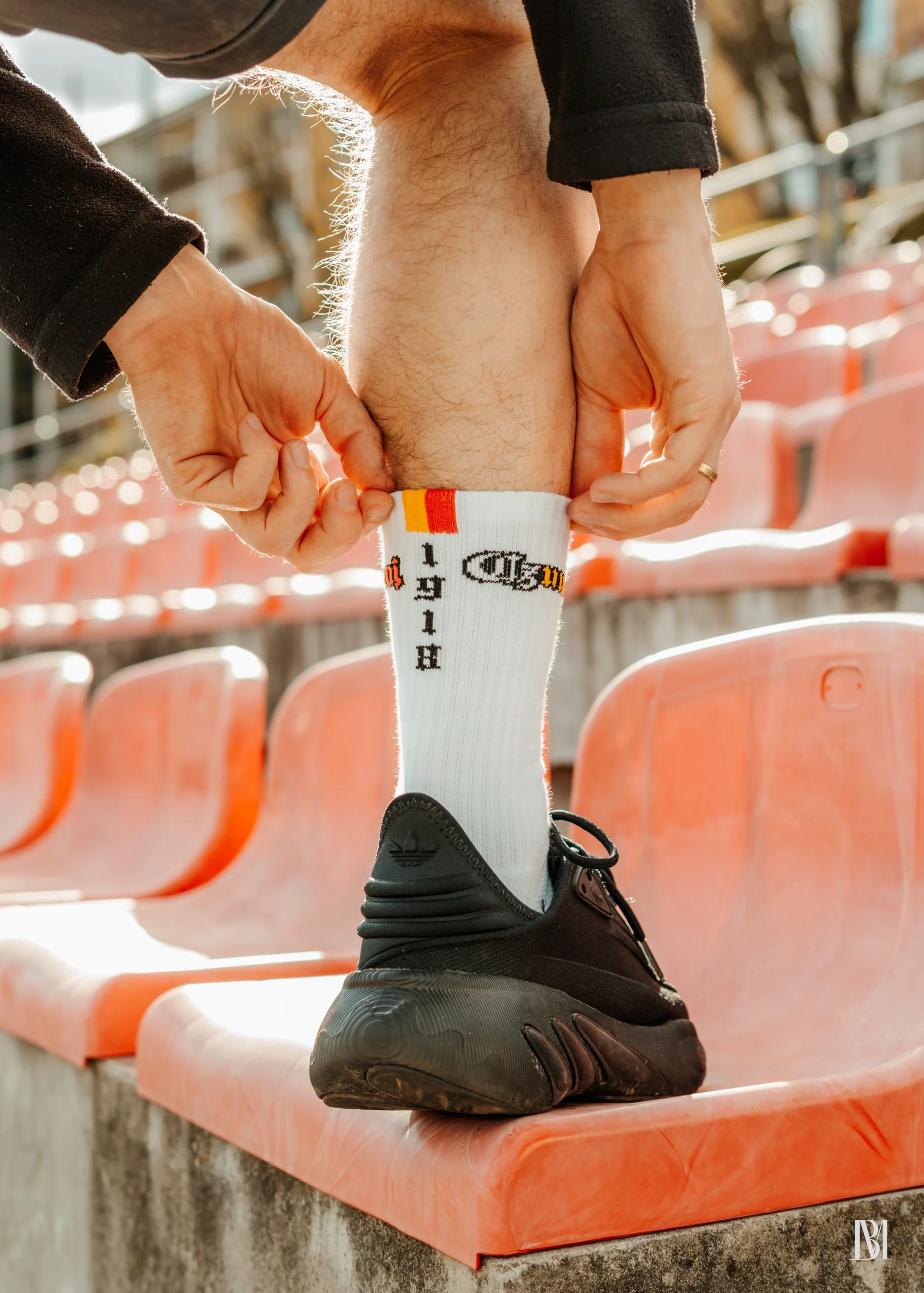 Białe skarpetki z mini flagą o kolorach biało-żółto-czerwonych i rokiem 1918 na tle stadionu, krzesełek i budynku klubowego.
